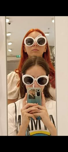 two women wearing sunglasses are looking at their cell phones in front of a mirror with the reflection of them