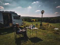 a woman sitting in a chair next to a camper on top of a lush green field