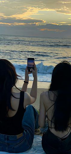 two women sitting on the beach taking pictures with their cell phones in front of them