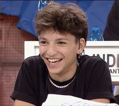 a young man sitting at a table with a smile on his face and wearing a black t - shirt