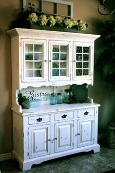 a white china cabinet with glass doors and flowers on the top, in a living room