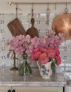 pink flowers are in vases on the kitchen counter next to utensils and cutting boards