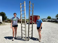 two women standing next to a ladder in the middle of a parking lot with trucks behind them