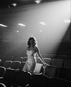 a woman in a white dress is standing on a stage with empty chairs and bright light coming from behind her