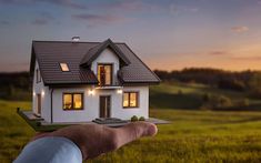 a hand holding a small model house in the middle of a field at sunset or dawn