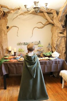 a little boy dressed in a green cape standing next to a table with food on it