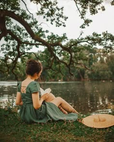 a woman sitting on the grass next to a body of water reading a book under a tree