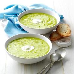 two white bowls filled with green soup next to bread and spoons on a table