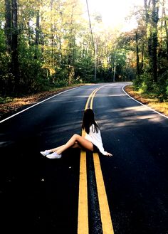 a woman sitting on the side of an empty road in the middle of the woods