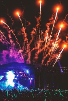 fireworks are lit up in the night sky above an outdoor concert stage as people watch