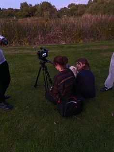 three people sitting in the grass while one person is recording something on a tripod