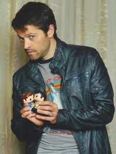 a man in a leather jacket is holding a toy and looking at the camera while standing next to a curtain