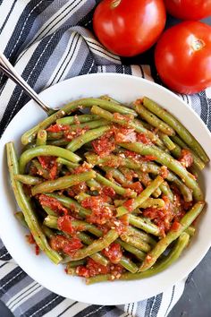 a white bowl filled with green beans covered in marinara sauce next to two tomatoes