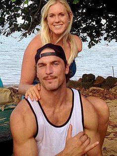 a man sitting on top of a woman's shoulders at the beach with trees in the background