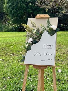 an easel with flowers on it in the grass next to a sign that says, welcome