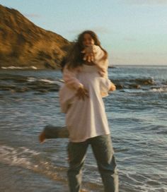 a woman walking on the beach with her dog in her arms and she's holding something up to her ear