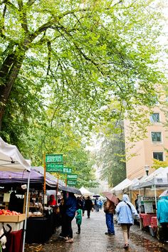 people walking through an open air market with umbrellas and tables full of goods under trees