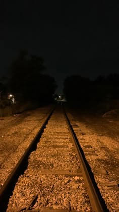 an empty train track at night with the lights on and trees in the distance behind it