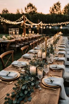 a long wooden table with candles and place settings on it, surrounded by greenery