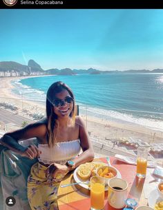 a woman sitting at an outdoor table with food and drinks on it, overlooking the beach