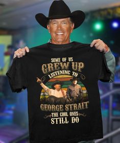 a man wearing a cowboy hat holding up a t - shirt that says, some of us grew up listening to george strait the cool ones still do