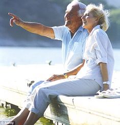 an older man and woman sitting on a dock pointing at something in the air with their hands