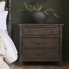 a nightstand with two drawers and a plant on it next to a bed in a bedroom