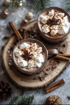 two mugs of hot chocolate with whipped cream and cinnamon on a wooden board surrounded by christmas decorations