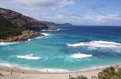 people are walking on the beach near the water and cliffs in the distance, with waves crashing onto the shore