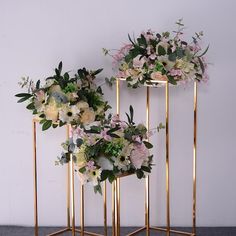 three gold plant stands with flowers and greenery on them, against a white wall