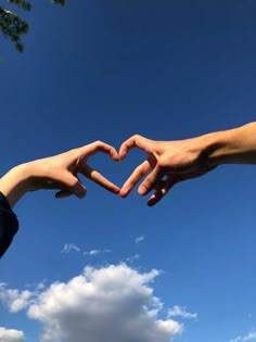 two hands making a heart shape with their fingers against a blue sky and white clouds