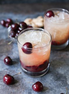 two glasses filled with ice and cherries on top of a gray table next to nuts