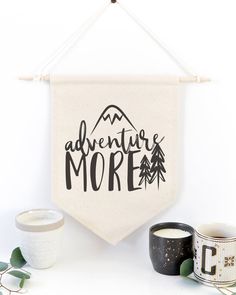 an adventure more banner hanging on a wall next to two mugs and some plants