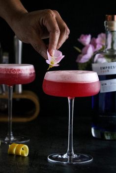 a person placing a flower on the top of a pink drink in two coupe glasses