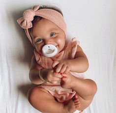 a baby with a pacifier in her mouth laying on a white sheet wearing a pink outfit