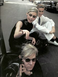 two men and a woman sitting on the ground in front of some cars, one with sunglasses on her head