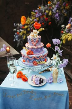 a blue table topped with a multi layer cake and wine glasses next to flowers in vases