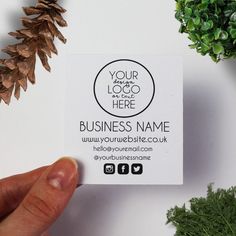 a person holding up a business card next to some pine cones and greenery on a white surface