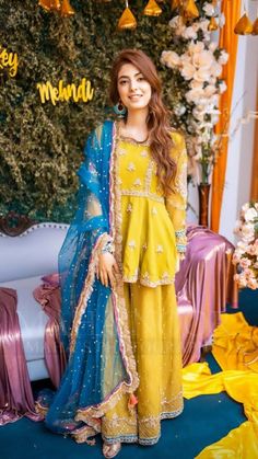 a woman in yellow and blue outfit standing next to a floral wall with flowers on it