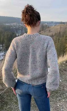 a woman standing on top of a hill wearing a knitted sweater and jeans with trees in the background