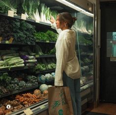 a woman is looking at vegetables in a grocery store while holding a shopping bag with her right hand