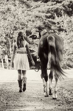 black and white photograph of a woman leading a horse down a dirt road with trees in the background