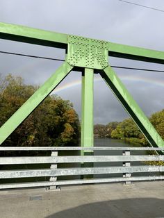 a green metal bridge with a rainbow in the sky and trees behind it, on a cloudy day