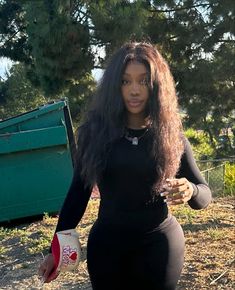 a woman with long hair in a black dress holding a drink and standing next to a dumpster