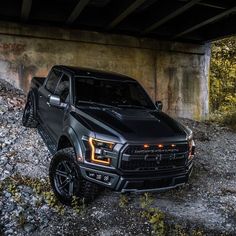 a black truck parked under a bridge on some rocks and grass with trees in the background