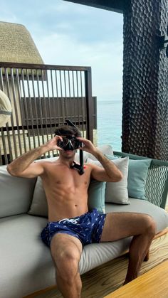 a shirtless man sitting on top of a couch next to the ocean holding a camera