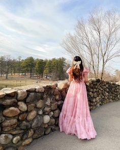 a woman in a pink dress leaning against a stone wall with her back to the camera