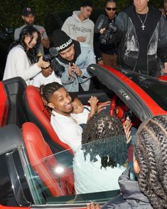 a group of people sitting around a red sports car with the hood up and one person waving