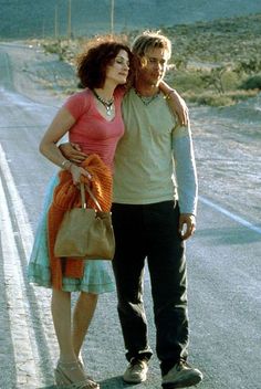 a man and woman standing in the middle of an empty road with mountains in the background