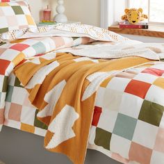 a bed covered in colorful checkered sheets and blankets next to a teddy bear on a window sill